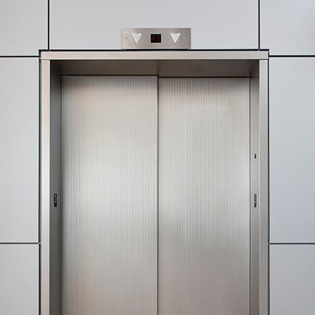 Closed stainless steel elevator doors set in a modern, light gray tiled wall, with a control panel above displaying floor information and up and down arrow buttons.
