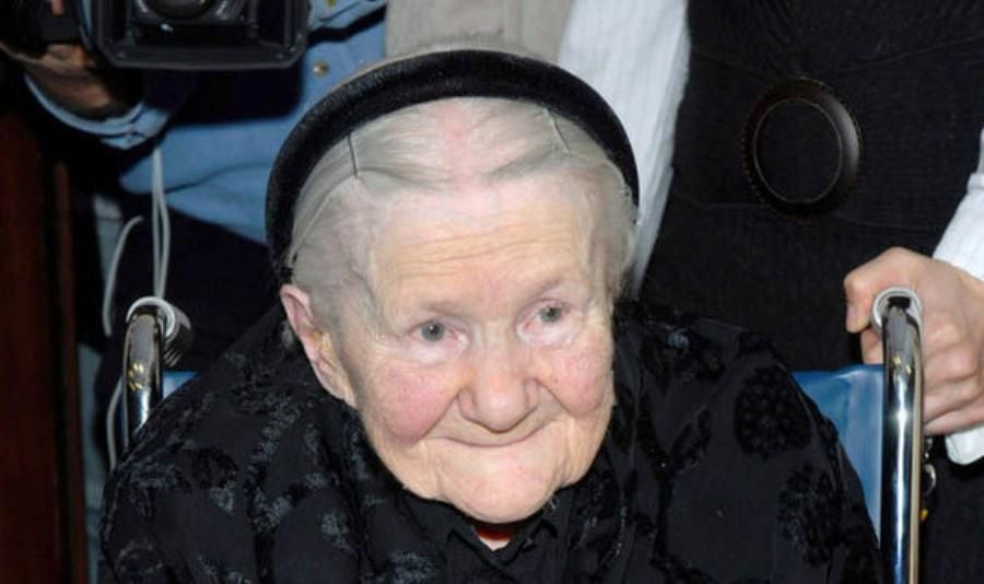 An elderly woman with short white hair, wearing a black headband and dark clothing, sits in a wheelchair and smiles gently. A person stands behind her, and part of a camera is visible in the background.