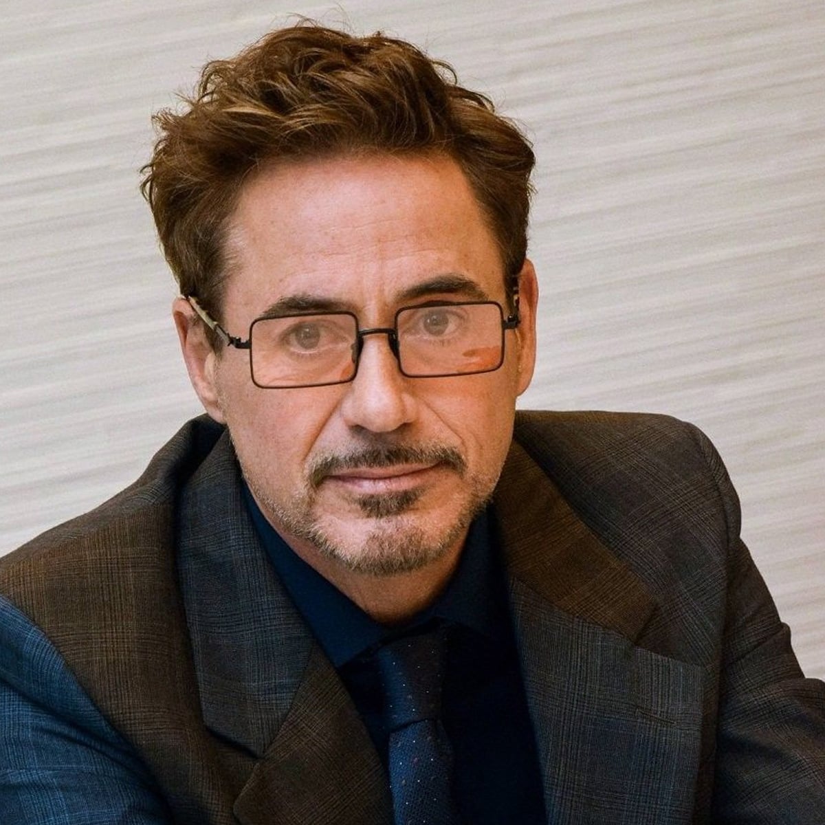 A man with short brown hair and a goatee wearing glasses, a dark suit jacket, shirt, and tie, sits against a light colored, textured background, looking directly at the camera.