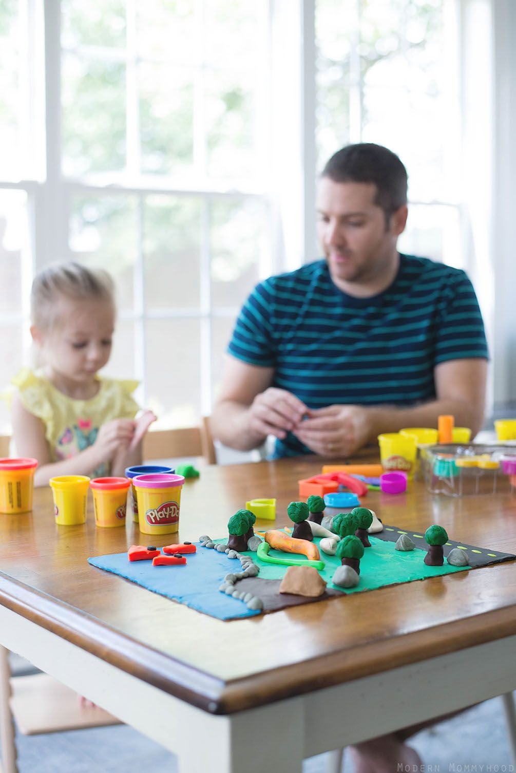 A man and a young girl sit at a table playing with colorful Play-Doh. Various containers and sculpted shapes, including trees and rocks, are spread across the table. Sunlight streams in through large windows behind them.
