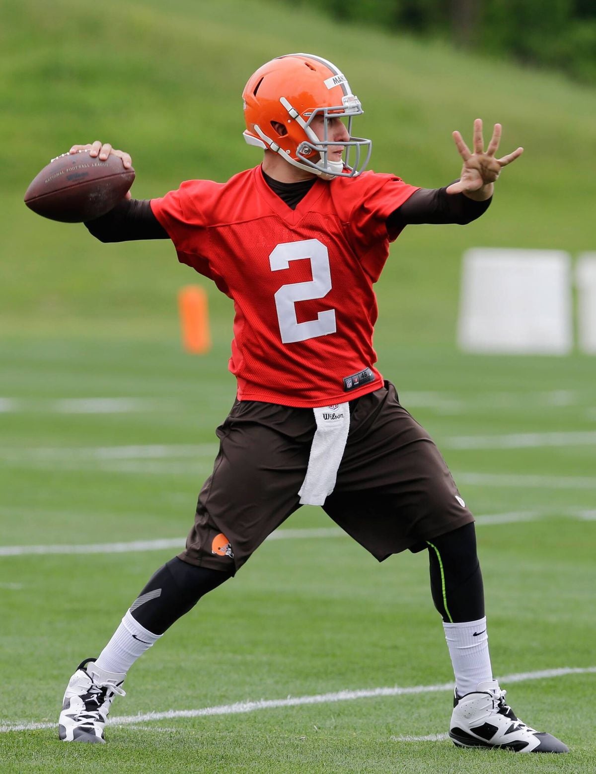 A football player in a red jersey, brown pants, and orange helmet is preparing to throw a football on a grassy field during practice.