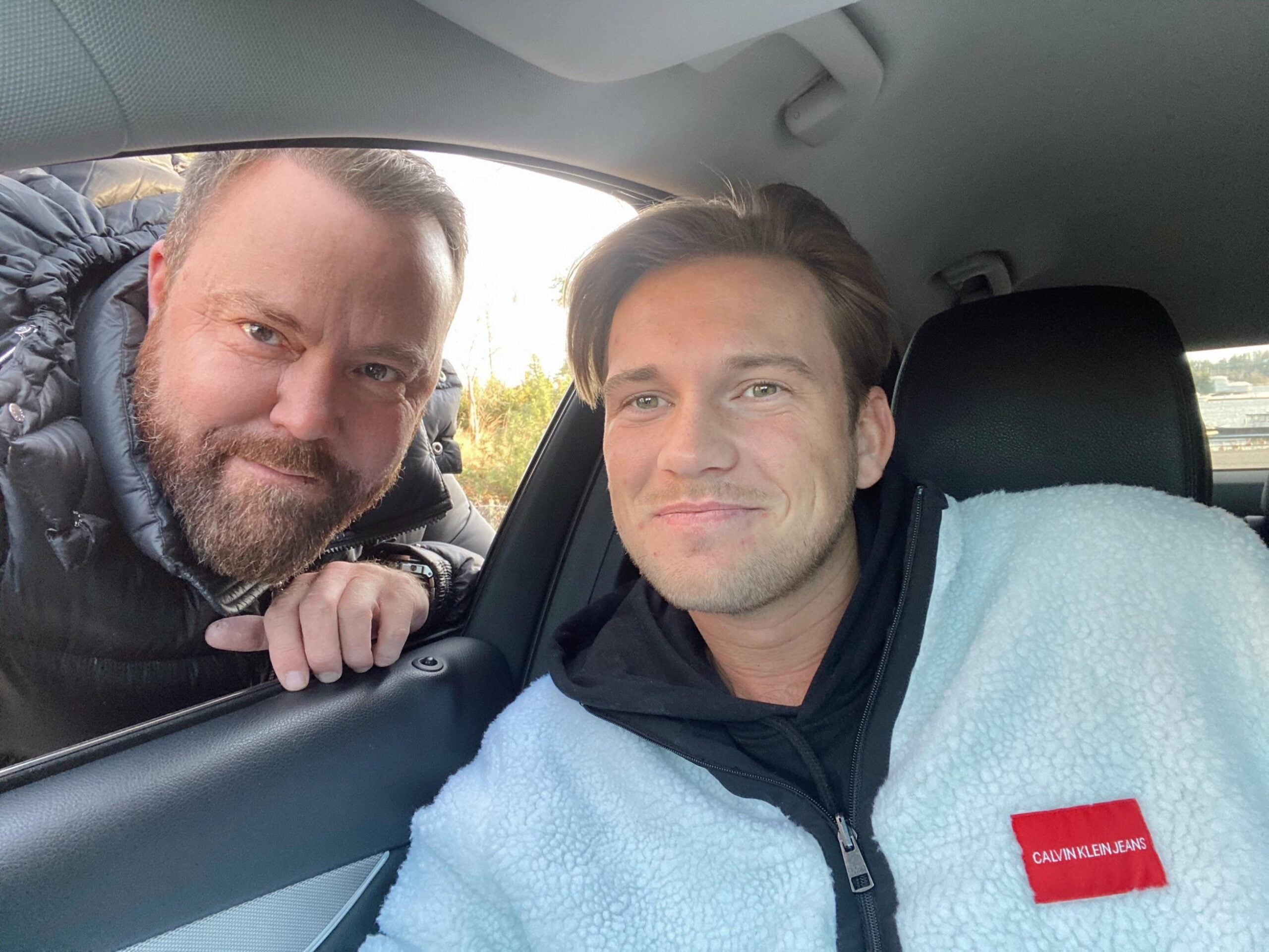 Two men smiling for a photo, one sitting inside a car wearing a white Calvin Klein fleece, and the other leaning in through the open window from outside, both appearing happy.