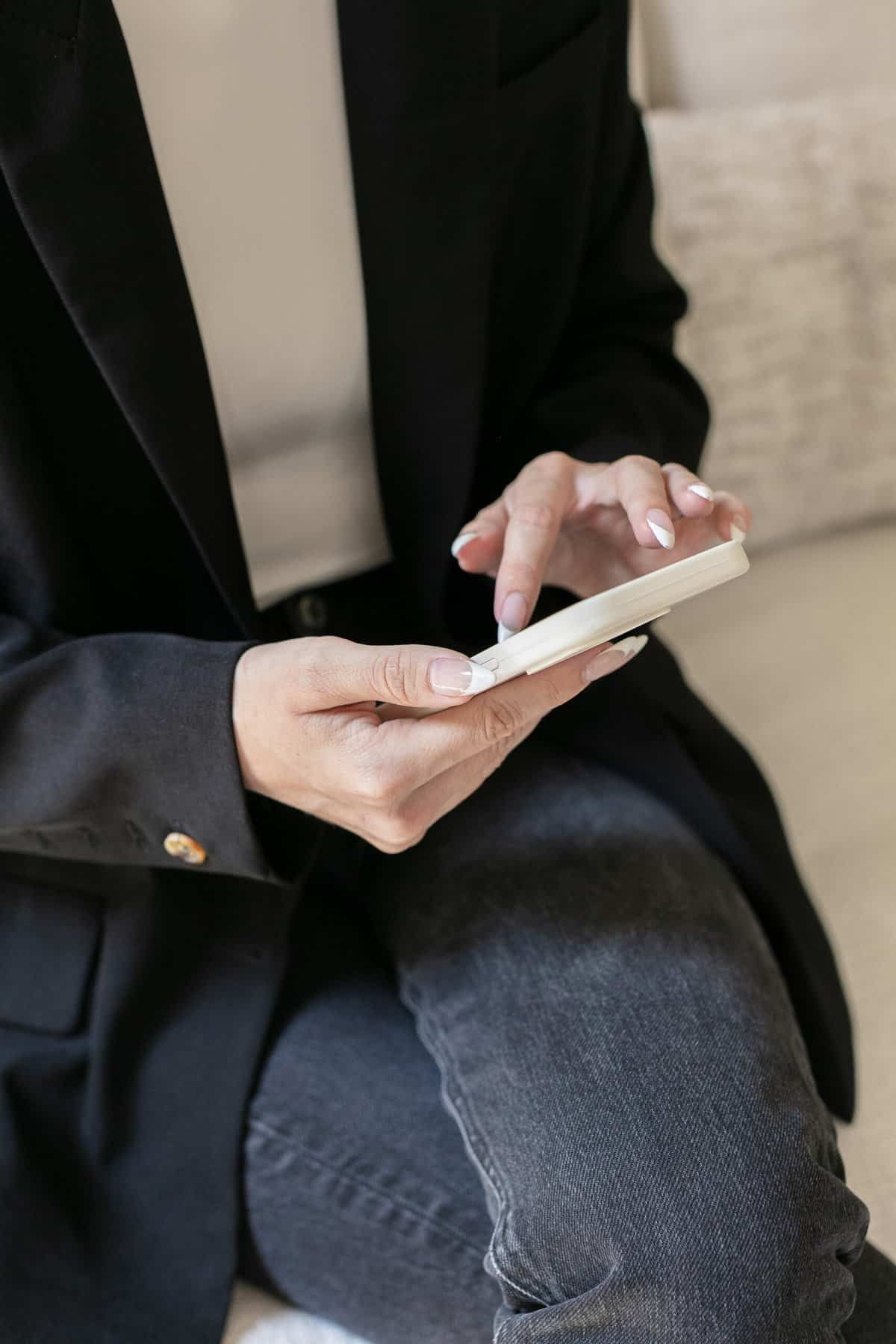 A person wearing a black blazer and gray jeans is sitting and using a smartphone, with their fingers interacting with the screen. The focus is on their hands and phone.