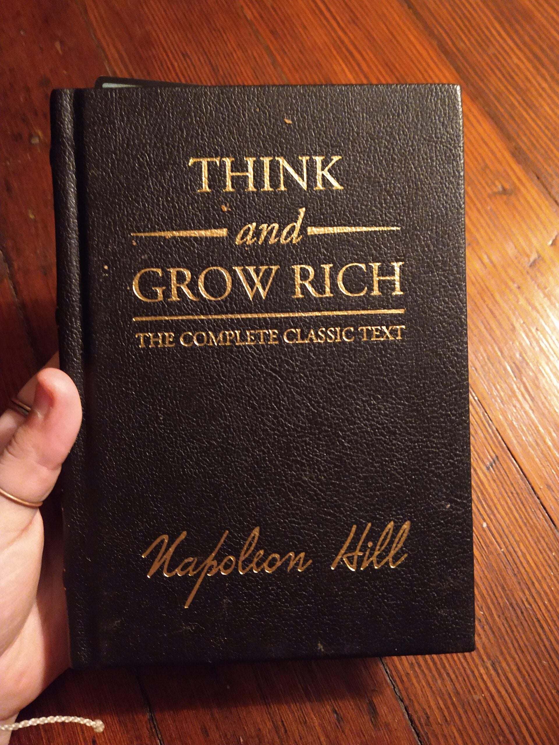 A hand holds a black hardcover book titled "Think and Grow Rich: The Complete Classic Text" by Napoleon Hill, with gold lettering on the cover. The background is a wooden surface.