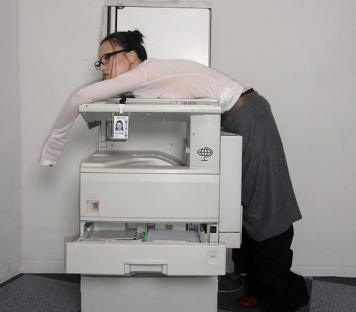 A woman wearing glasses and office clothes lies sprawled face down over a photocopier in a bare office, appearing exhausted or overwhelmed. Her ID badge hangs from her neck.