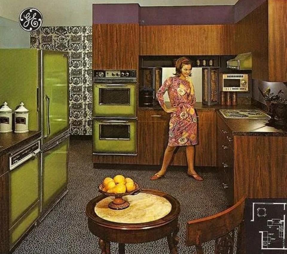 A woman in a colorful dress stands in a retro 1970s kitchen with green appliances, dark wood cabinets, patterned wallpaper, and a small round table with a bowl of fruit in the foreground.