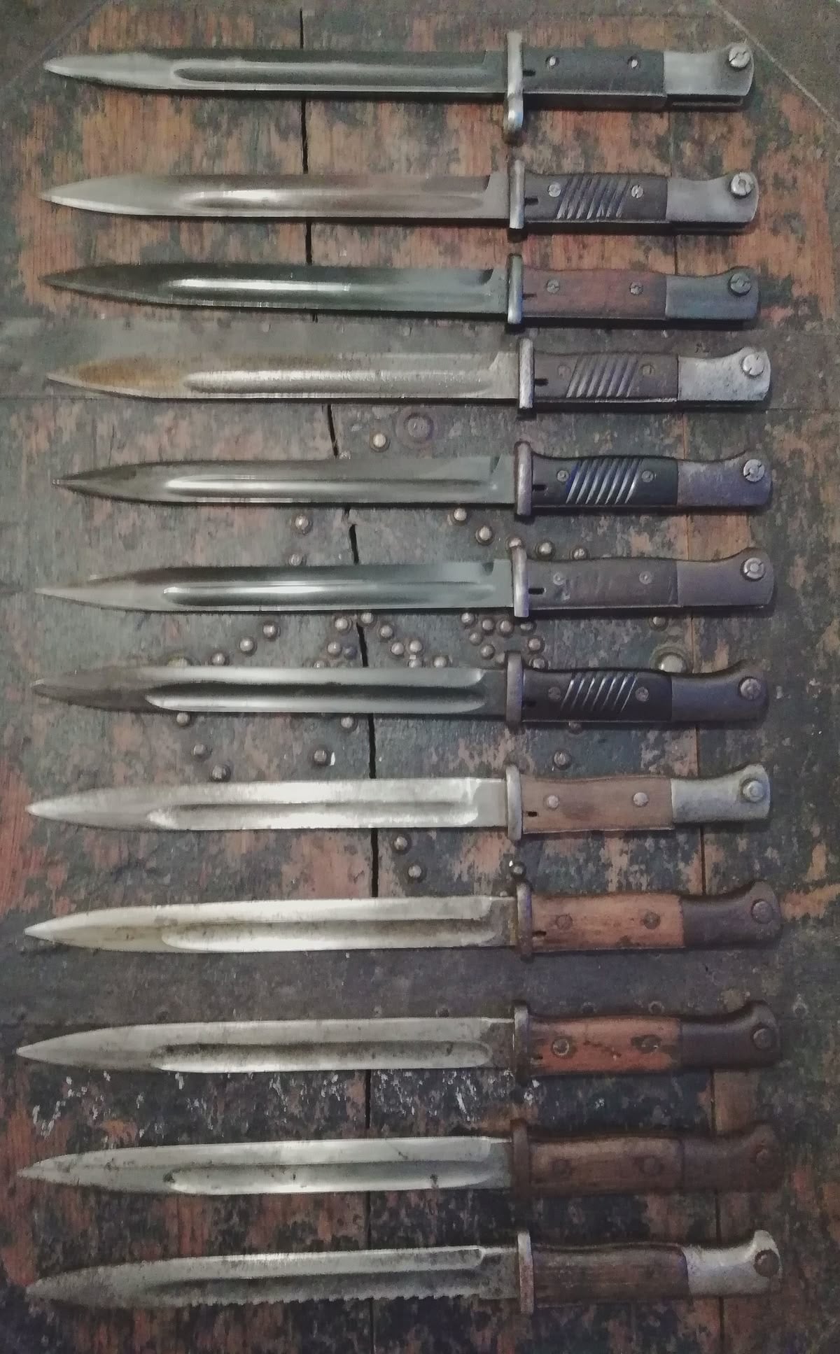 Twelve vintage bayonets with dark, aged handles are arranged in two neat rows on a worn wooden surface. The metal blades show signs of rust and wear, reflecting their age and use.