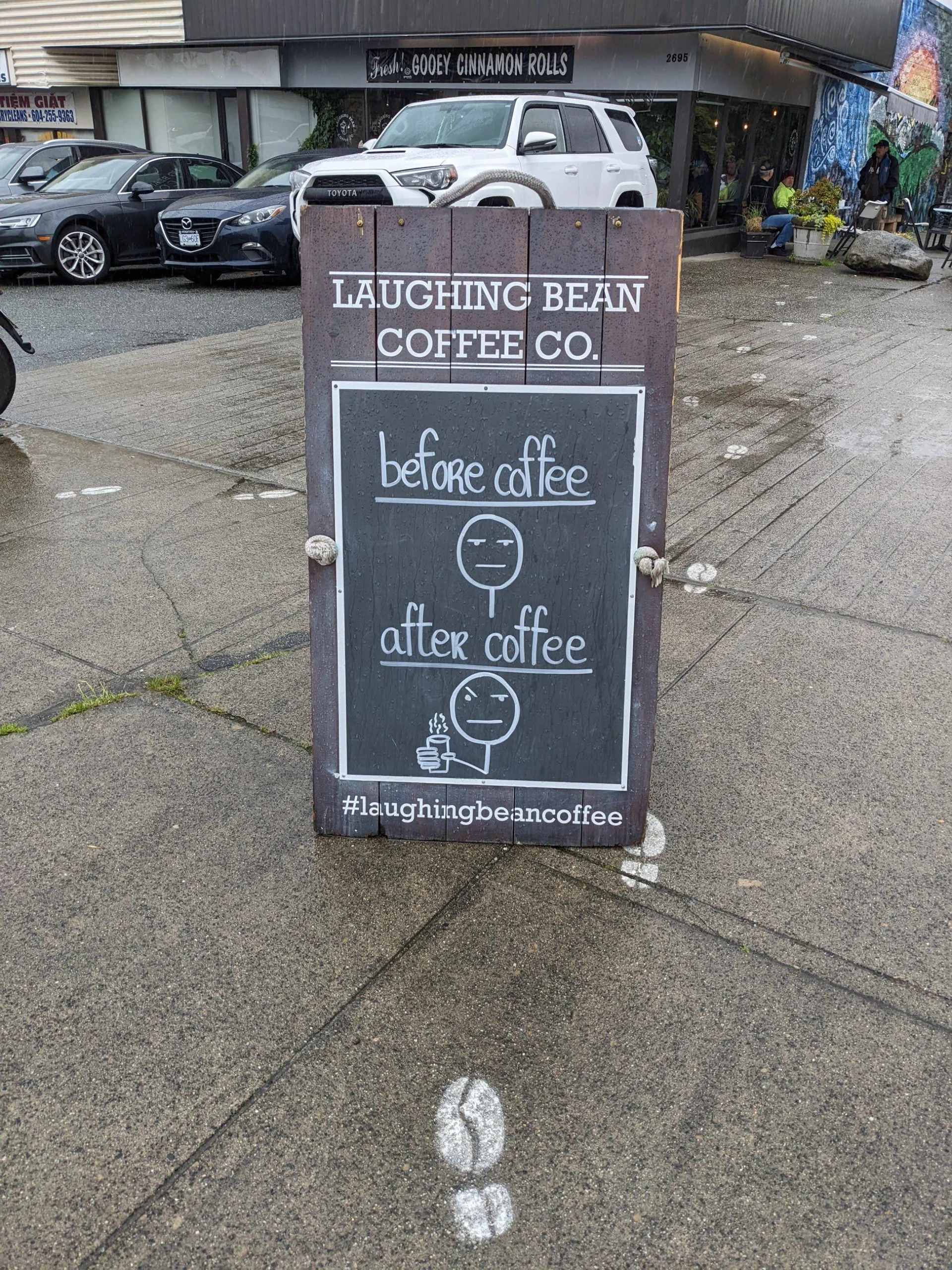 A sidewalk sign for Laughing Bean Coffee Co. shows a sad face labeled “before coffee” and a happy face holding a cup labeled “after coffee.” The sign includes the hashtag #laughingbeancoffee.