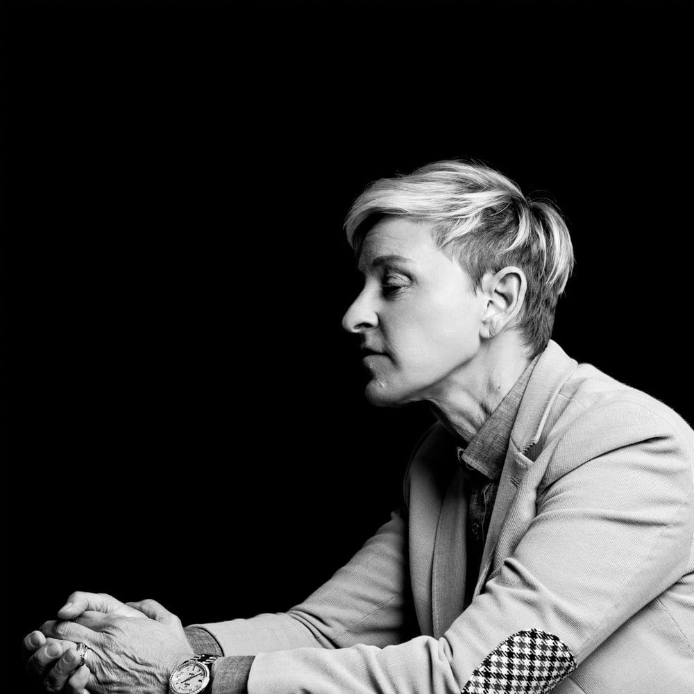 A person with short, light hair sits in profile against a black background, wearing a light-colored blazer with a checkered elbow patch and a watch, hands clasped in front of them.
