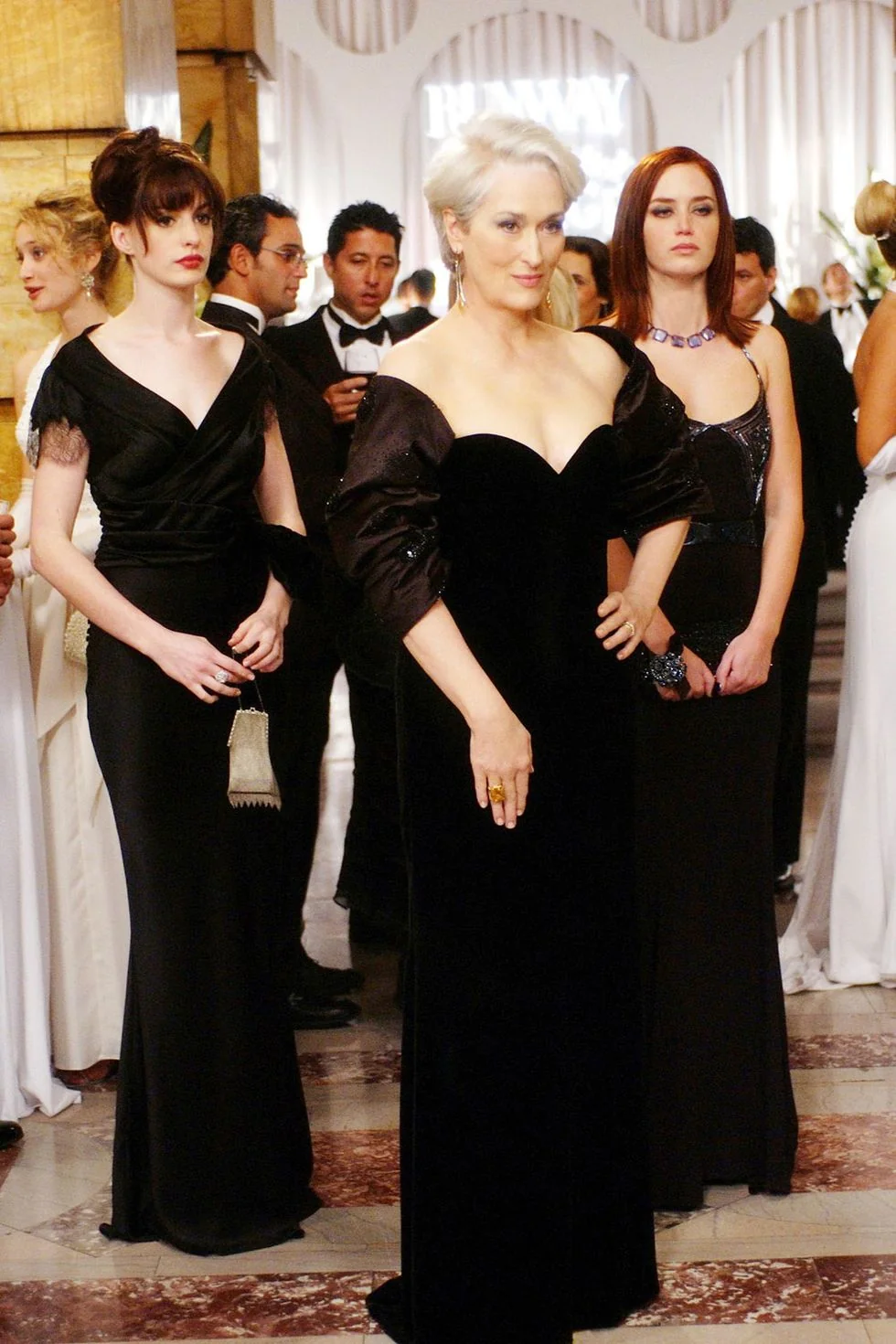Three women in elegant black evening gowns stand together at a glamorous, upscale event, surrounded by guests in formal attire. The central woman has short white hair and stands confidently with hands on hips.