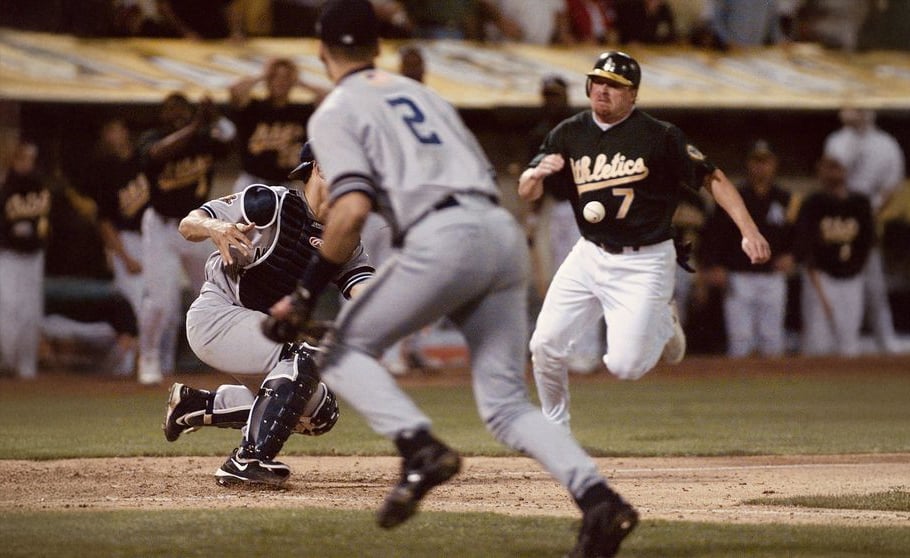 A baseball player in a green "Athletics" uniform runs toward home plate as the catcher attempts to tag a sliding opponent. Another player in a gray uniform runs toward the action. The background shows teammates watching.