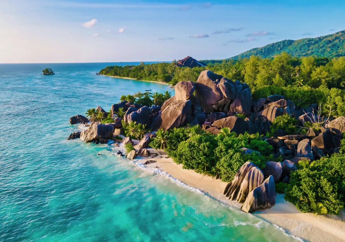Aerial view of a tropical beach with turquoise water, white sand, large granite boulders, and lush green trees. A forested hill is visible in the background under a blue sky with scattered clouds.