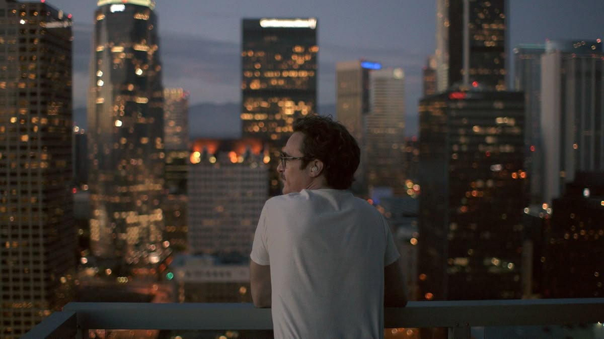 A person wearing glasses and a white t-shirt stands on a balcony at dusk, overlooking a city skyline with tall buildings and colorful lights.