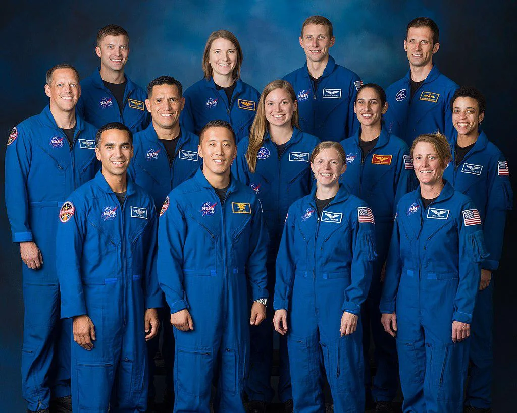 Thirteen astronauts wearing blue NASA flight suits stand in two rows against a blue backdrop, smiling and posing for a group photo. NASA and American flag patches are visible on their uniforms.