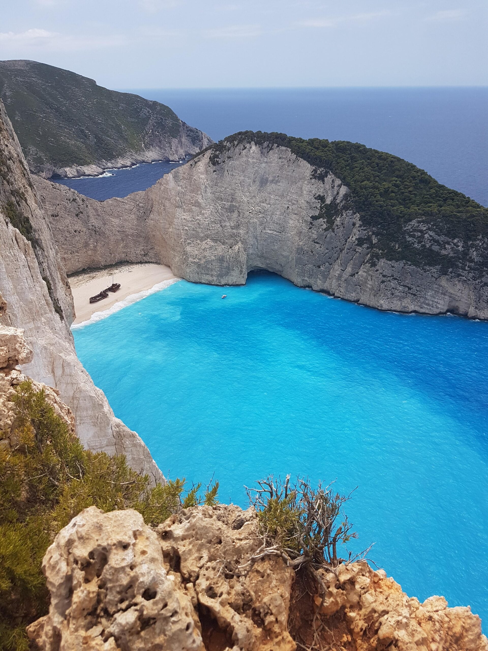 View of a secluded beach with turquoise water surrounded by steep cliffs and lush greenery; a shipwreck is visible on the white sand, with blue sea meeting the horizon under a clear sky.
