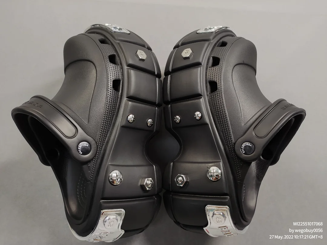 A pair of black Crocs with metallic accents, thick rubber soles, and metal screws, facing each other on a gray surface. The shoes appear heavily customized with an industrial design.