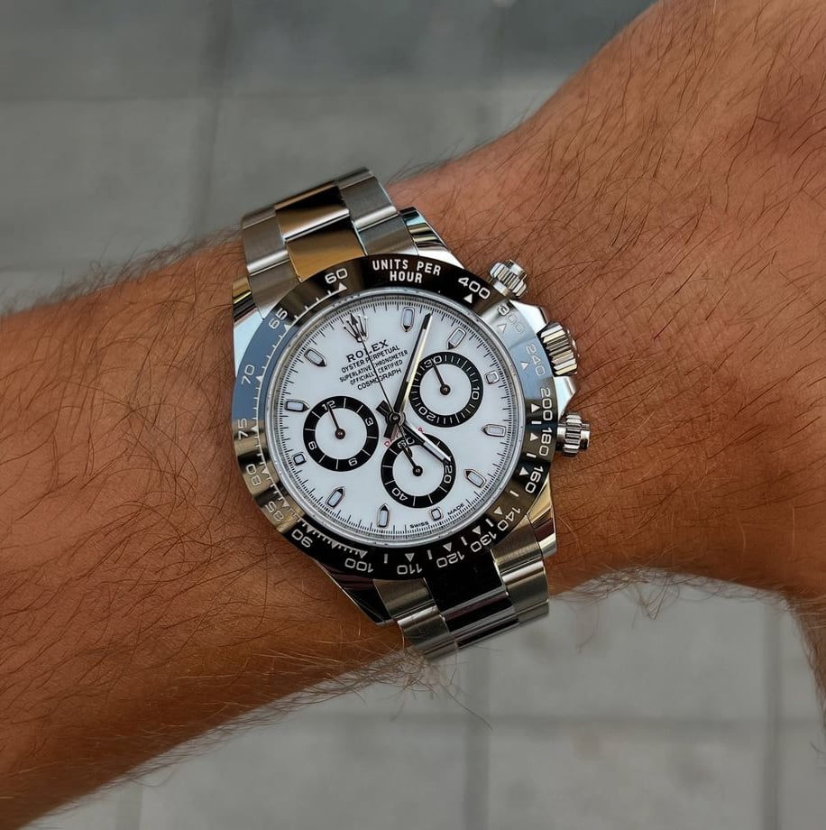 A close-up of a wrist wearing a stainless steel Rolex Daytona watch with a white dial and black ceramic bezel, featuring three subdials and silver hour markers. The background is blurred grey tiles.