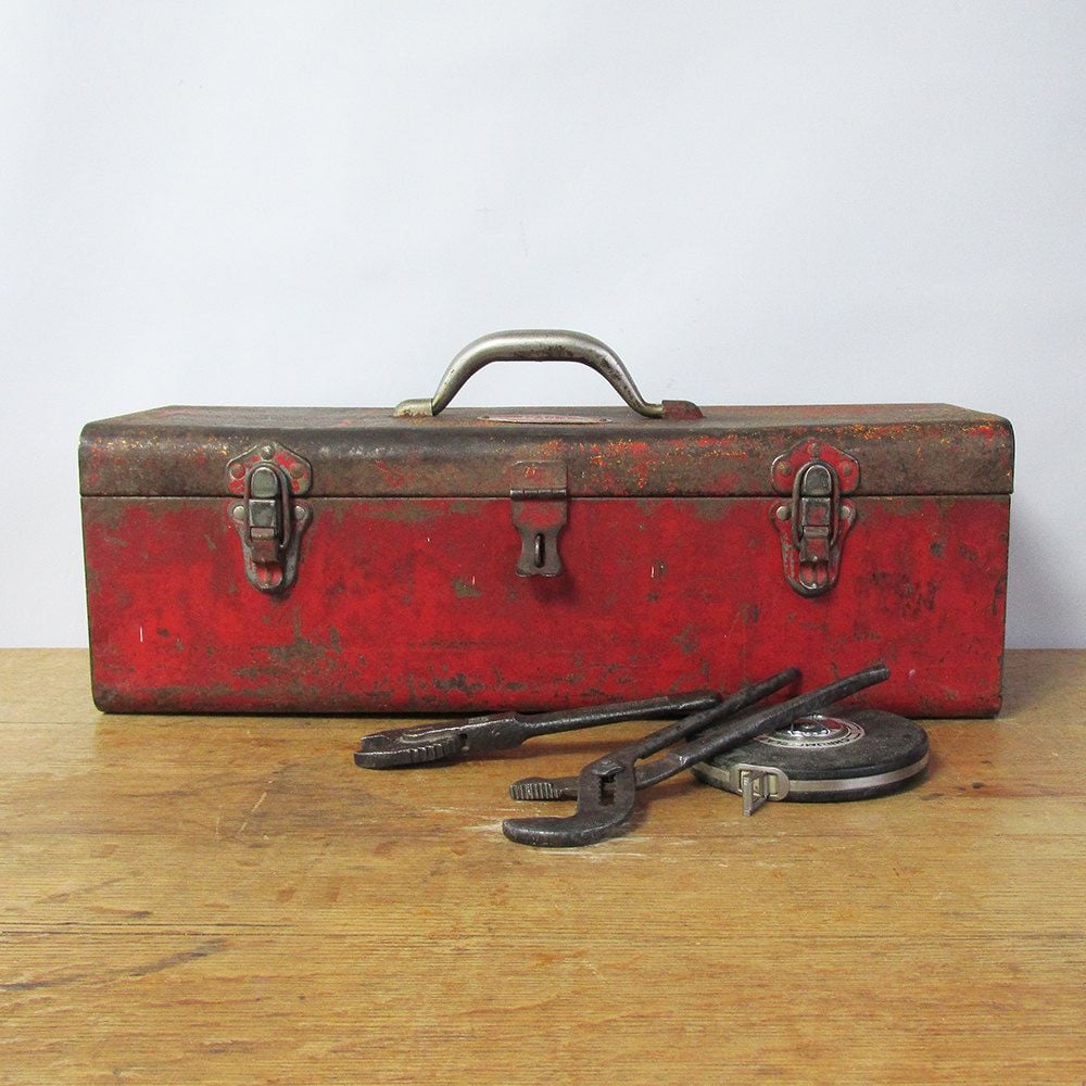 A rusty red metal toolbox with a handle sits on a wooden surface. In front of it are three old tools, including a wrench, pliers, and a tape measure, against a plain light background.