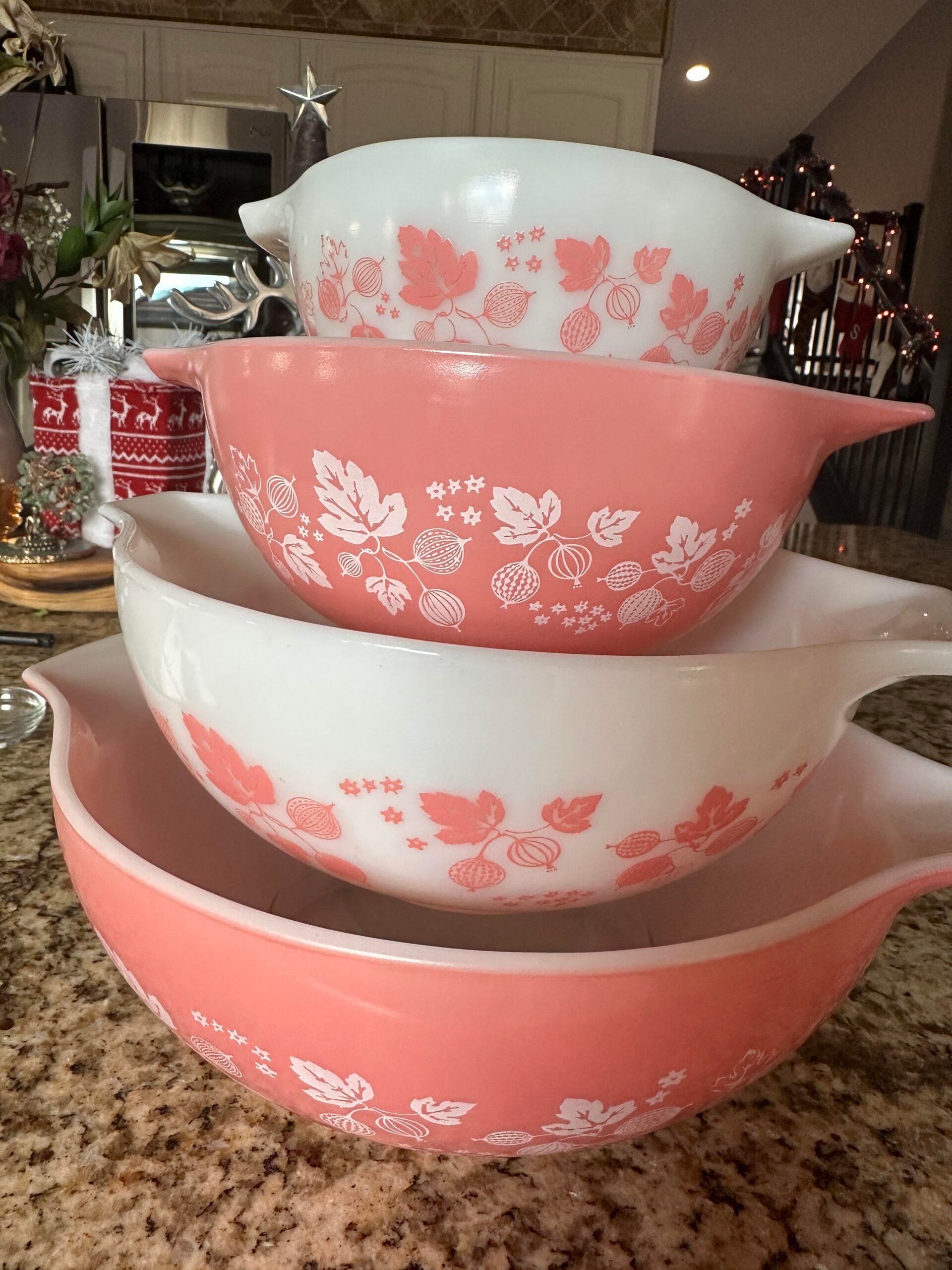 Four vintage mixing bowls with pink and white floral designs are stacked on a granite countertop in a kitchen with holiday decorations in the background.
