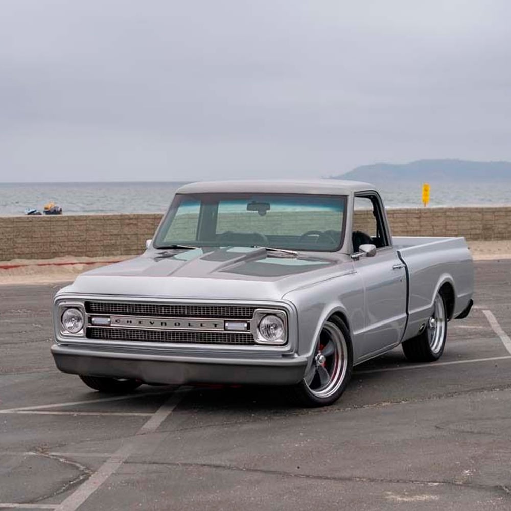 A restored silver Chevrolet pickup truck is parked in an empty lot near a beach on a cloudy day, with the ocean and a low brick wall visible in the background.