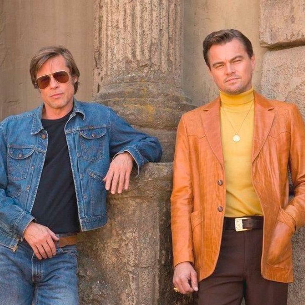 Two men stand side by side against a stone wall. One wears a denim jacket and sunglasses, while the other wears a tan leather jacket over a yellow turtleneck, looking confidently at the camera.