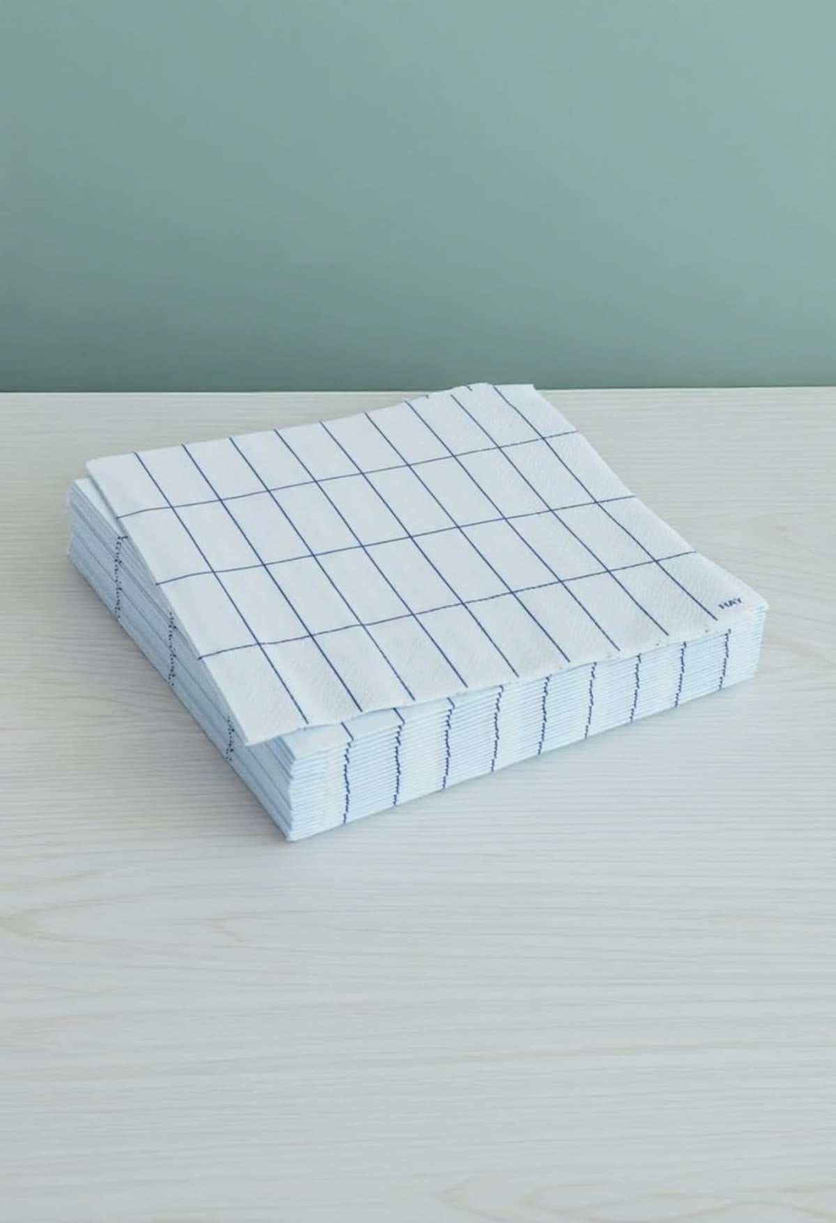 A stack of neatly folded white dish towels with a simple blue grid pattern sits on a light wooden surface against a muted green background.