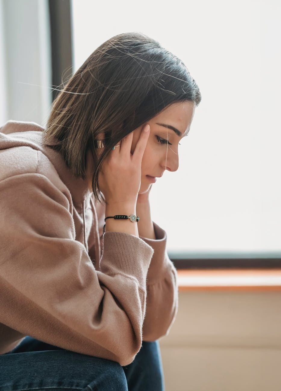 A young woman with short brown hair, wearing a beige hoodie and a bracelet, sits indoors looking down with her hands on her temples, appearing thoughtful or stressed.