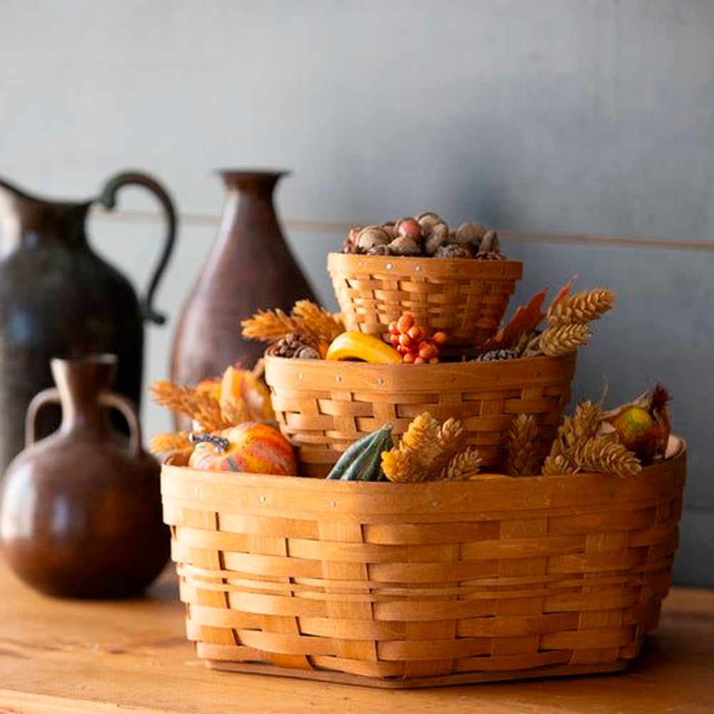 Three stacked woven baskets filled with autumn decorations like gourds, wheat stalks, and acorns sit on a wooden table, with three ceramic jugs in the background against a gray wall.