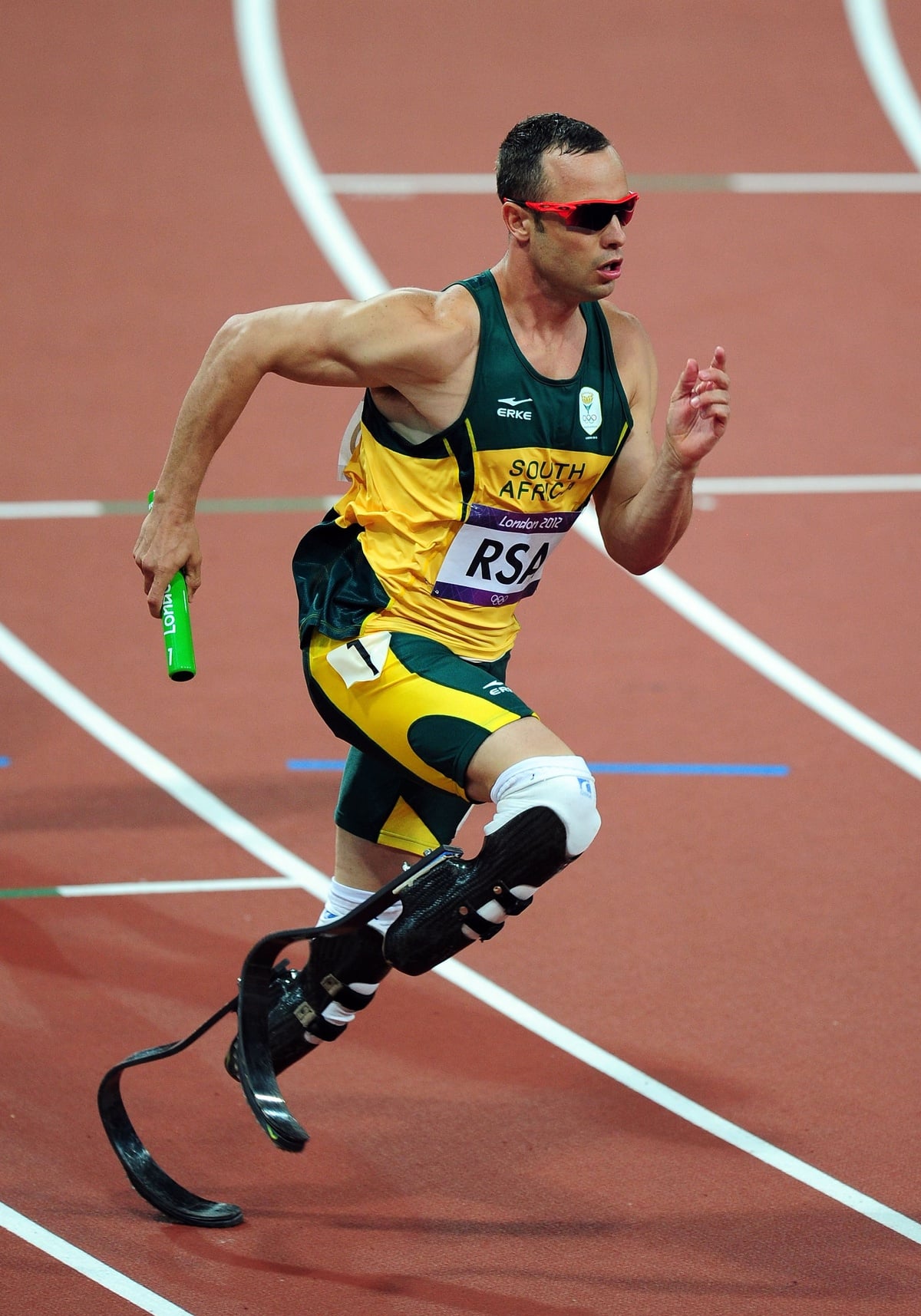 A male Paralympic sprinter from South Africa runs on a track with prosthetic blade legs, wearing a green and yellow uniform and red sunglasses, while holding a green relay baton.