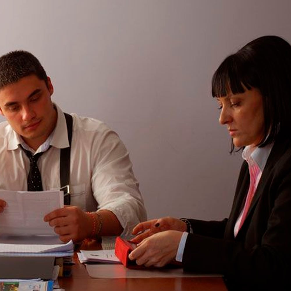 Two people in business attire sit at a table, looking at documents and a red object. One person is reviewing papers, while the other appears to be examining the red item. The scene suggests a professional or office setting.