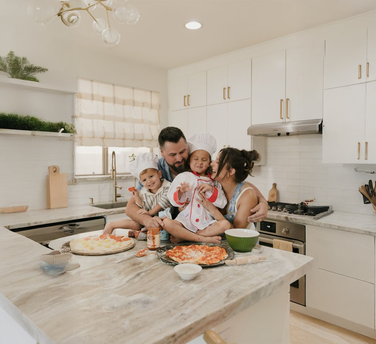 A family of four, wearing chef hats and aprons, sits together on a kitchen counter, smiling and hugging while making pizzas. Pizza ingredients and utensils are spread out on the countertop.