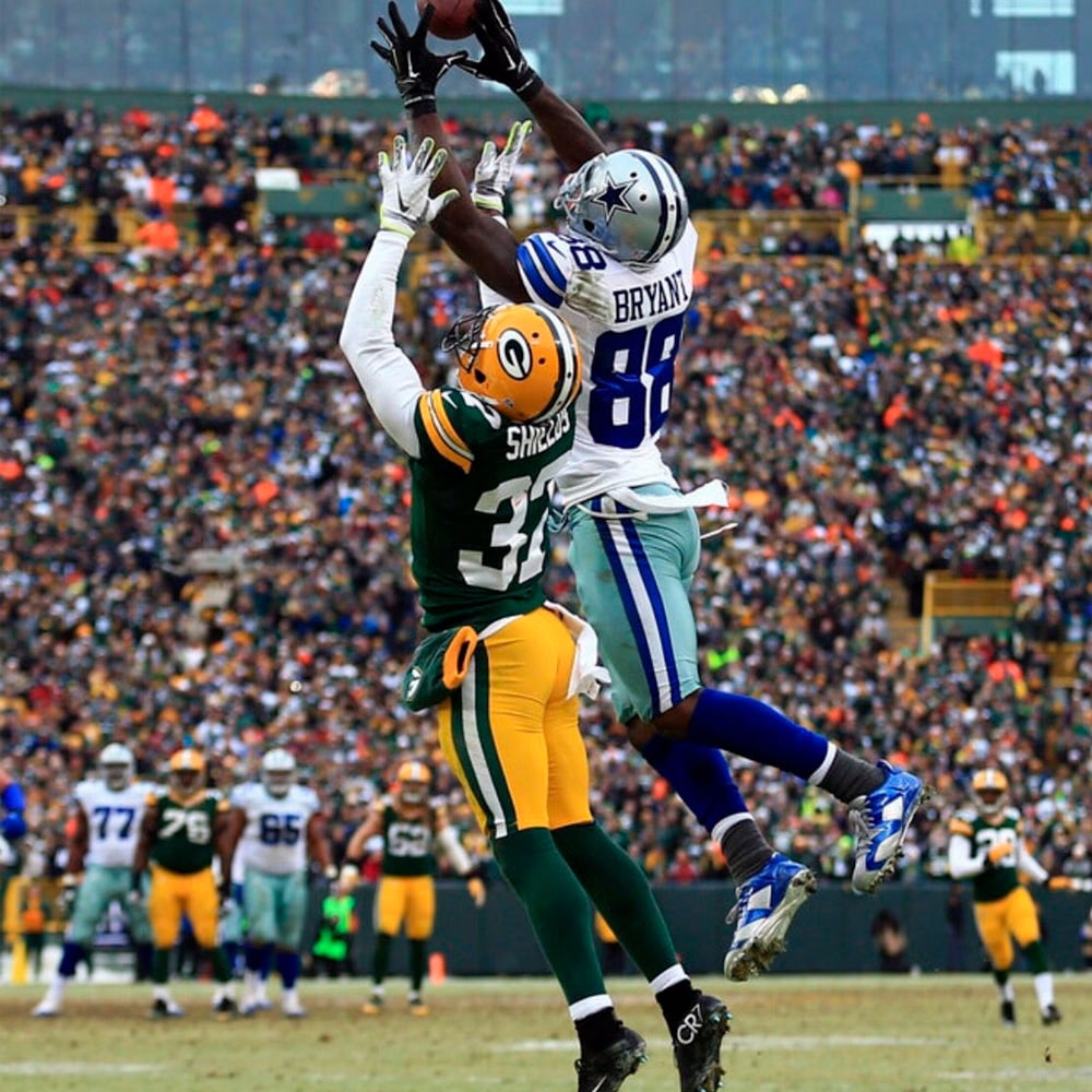 A Dallas Cowboys player jumps to catch a football mid-air while a Green Bay Packers defender tries to block him during an NFL game, with a crowd of fans in the background.