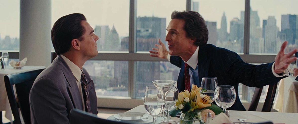 Two men in suits sit at a restaurant table with flower centerpiece and glassware, one speaking animatedly with arms outstretched; city skyline visible through large windows behind them.