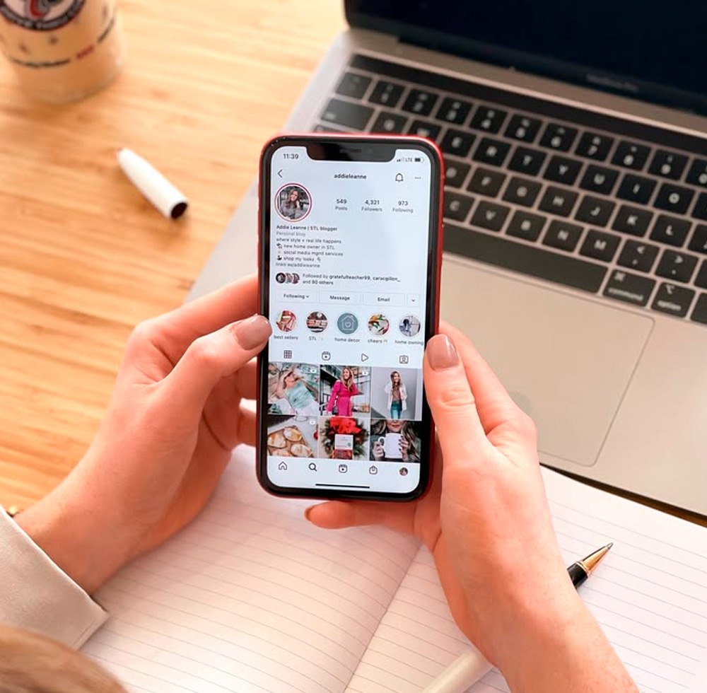A person holds a smartphone displaying an Instagram profile while sitting at a desk with a laptop, a notebook, a pen, and a drink. The person's hand is partially visible, and the workspace appears organized.