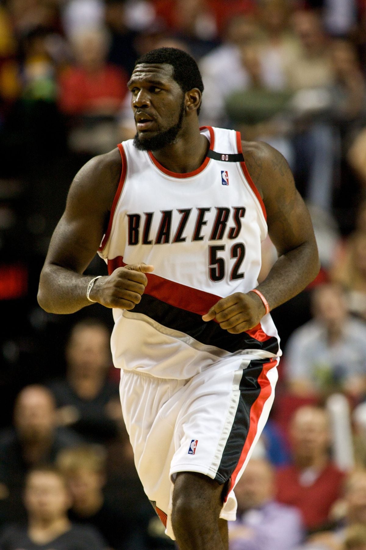 A basketball player in a white Portland Trail Blazers uniform with the number 52 runs on the court during a game. The background shows a blurred crowd of spectators.