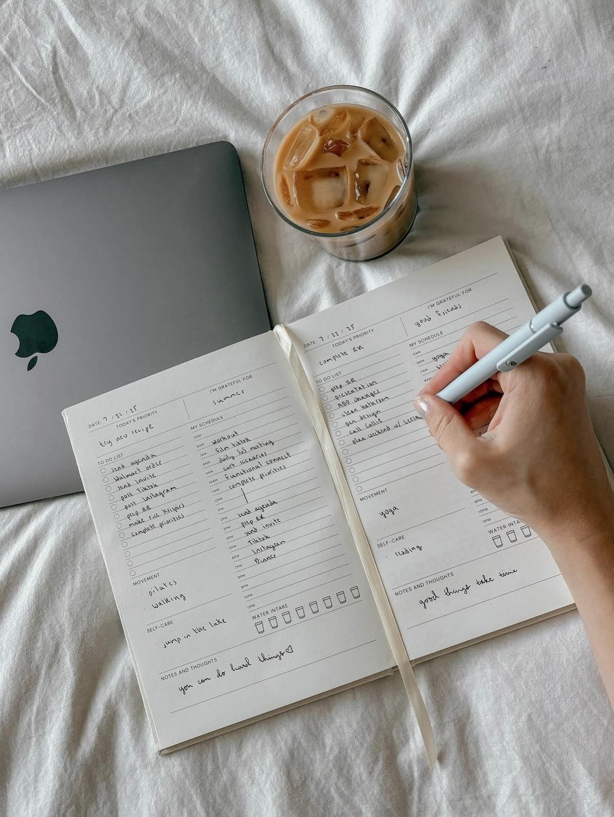 A hand writes in an open planner on a bed next to a closed MacBook and a glass of iced coffee. The planner contains to-do lists, notes, and checkboxes.