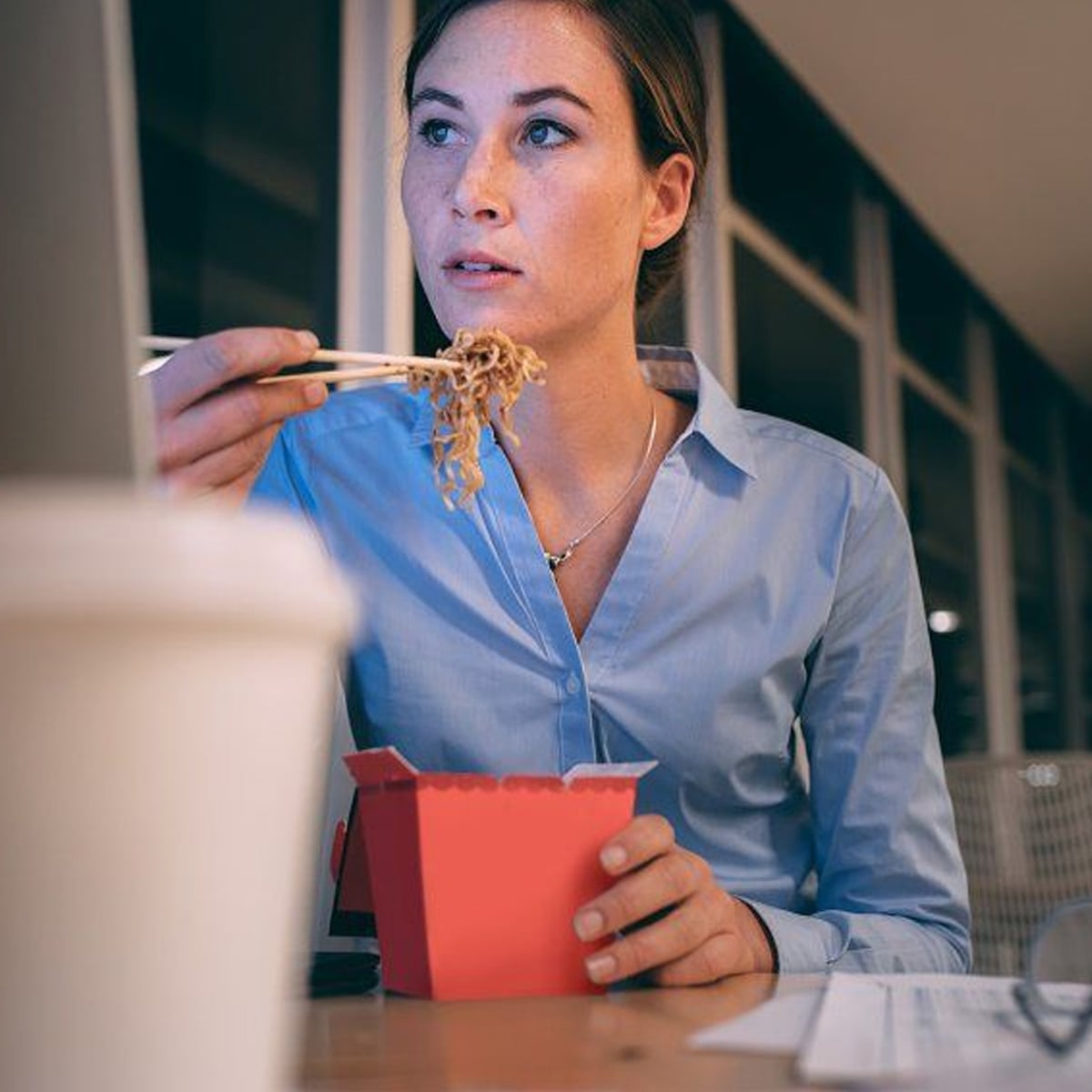 A woman in a blue shirt eating noodles with chopsticks from a red takeout box while sitting at a desk, appearing focused and working late in an office with large windows.