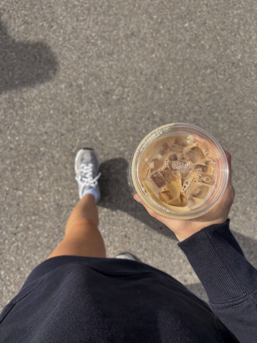 A person in a black sweatshirt and white sneakers walks on pavement, holding a clear plastic cup of iced coffee with milk, viewed from above.
