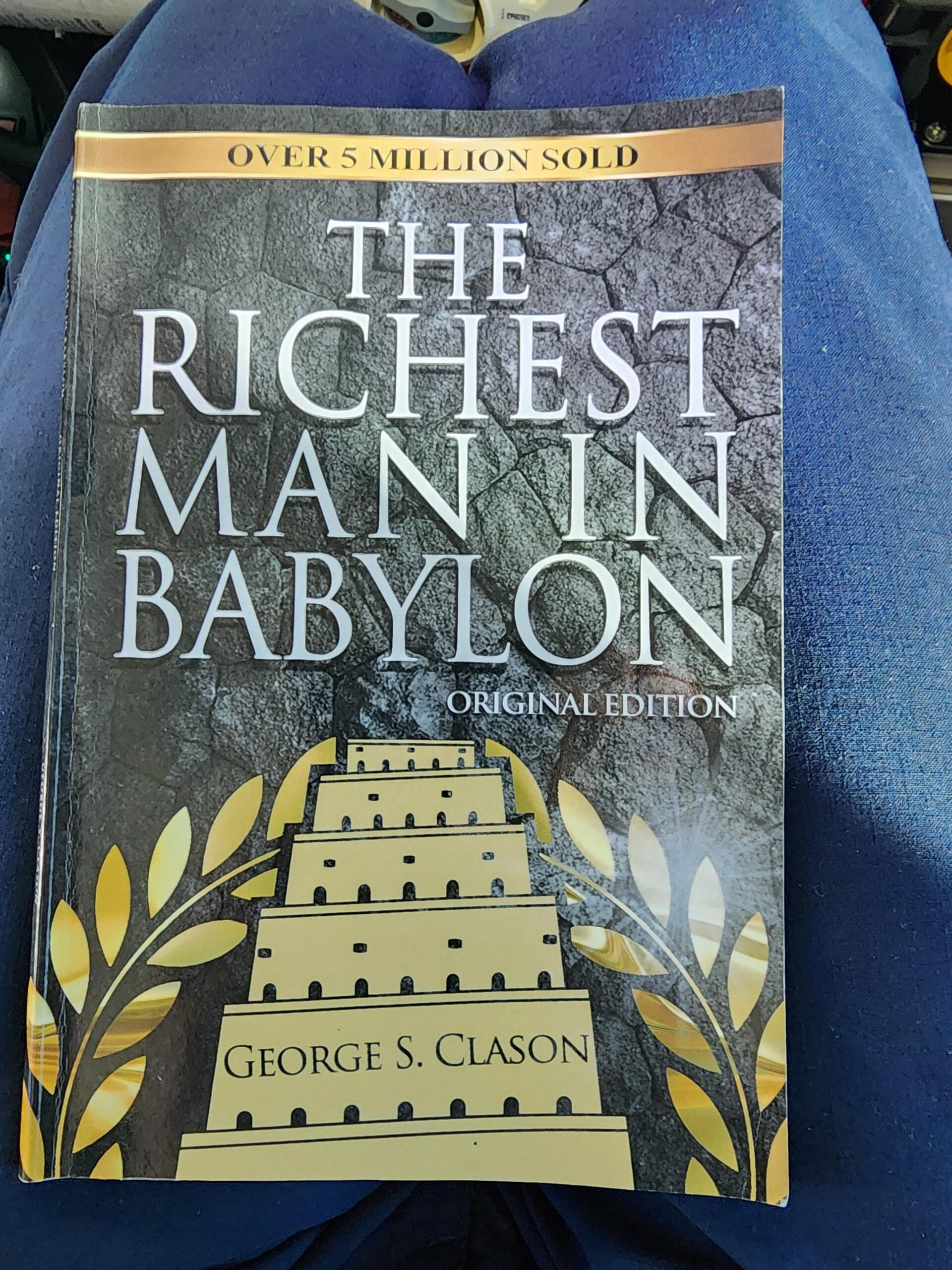 A person holding the book "The Richest Man in Babylon" by George S. Clason, featuring a golden ziggurat and laurel branches on the cover, with the text "Over 5 Million Sold" at the top.