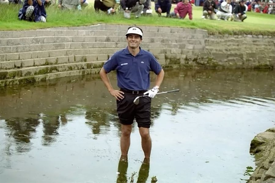 A golfer stands knee-deep in a water hazard, smiling and holding a golf club, while spectators watch from grassy steps in the background.