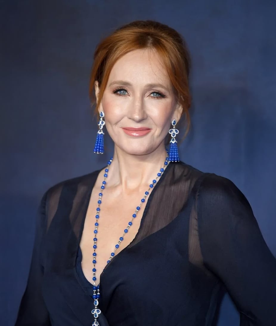 A woman with red hair in an updo wearing long blue earrings and a matching blue beaded necklace, dressed in a dark sheer-sleeved top, smiles in front of a dark blue background.