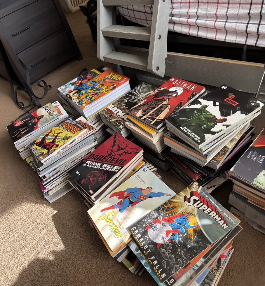 Stacks of comic books, including Batman, Superman, Daredevil, X-Men, and others, spread out on a carpeted floor near a bed and nightstand in a sunlit room.