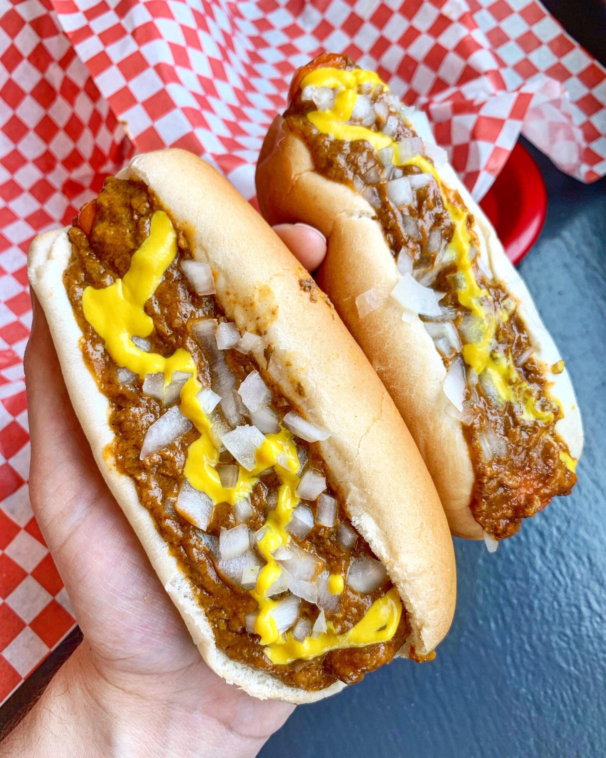 A hand holds two chili dogs topped with chopped onions and mustard, with red and white checkered paper lining in the background.