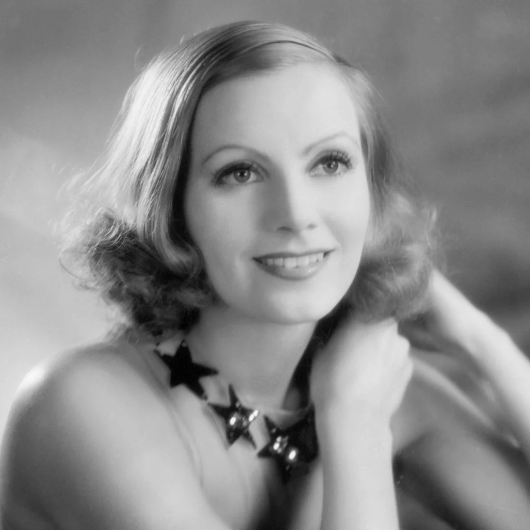 A woman with wavy, shoulder-length hair smiles softly, looking upward. She wears a star-shaped necklace and rests her arms near her neck. The image is black and white with a soft, classic Hollywood glamour style.