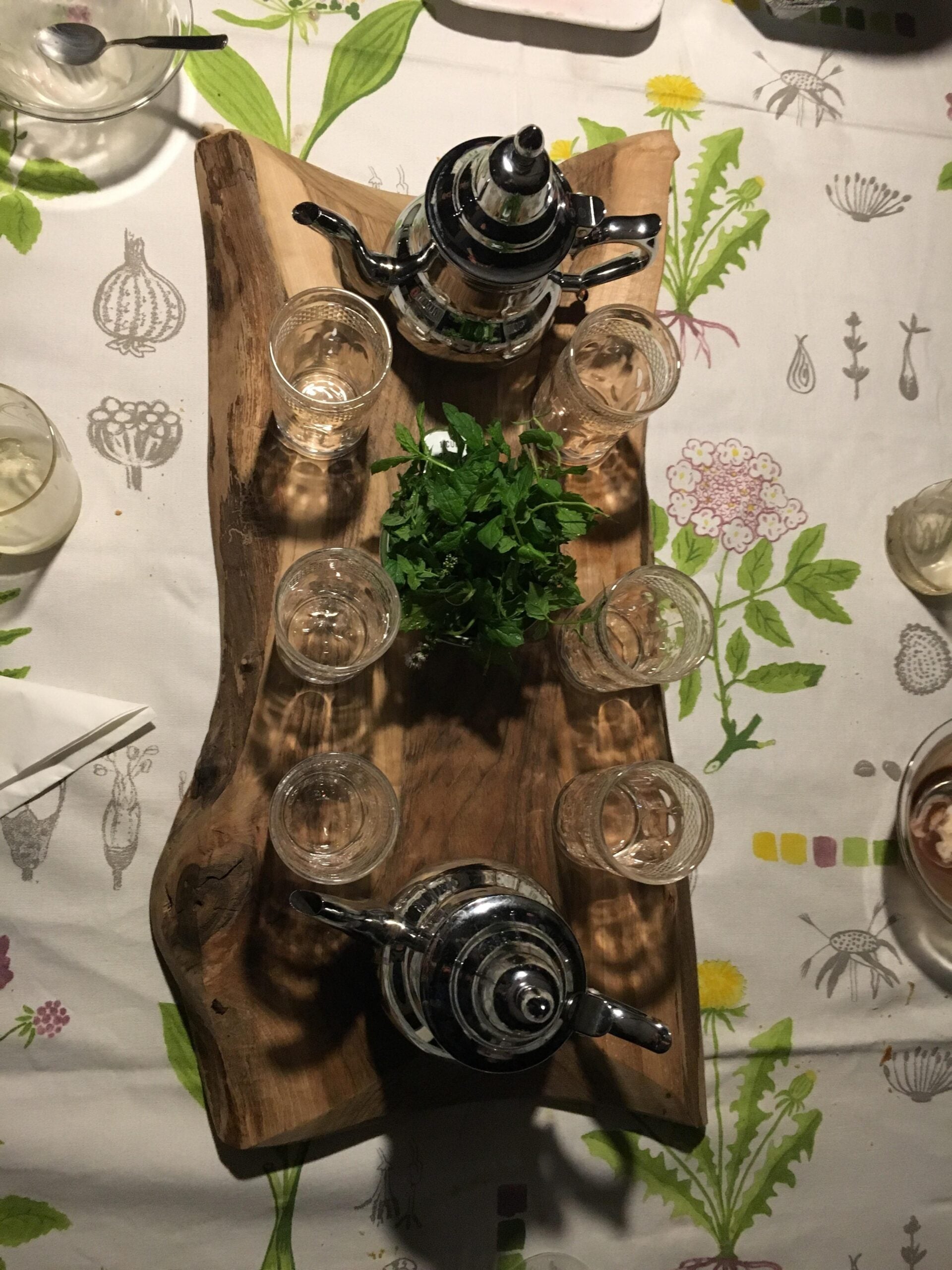 A wooden tray with two metal teapots, six clear glass tea cups, and a bunch of fresh mint on a table covered with a floral-patterned tablecloth.
