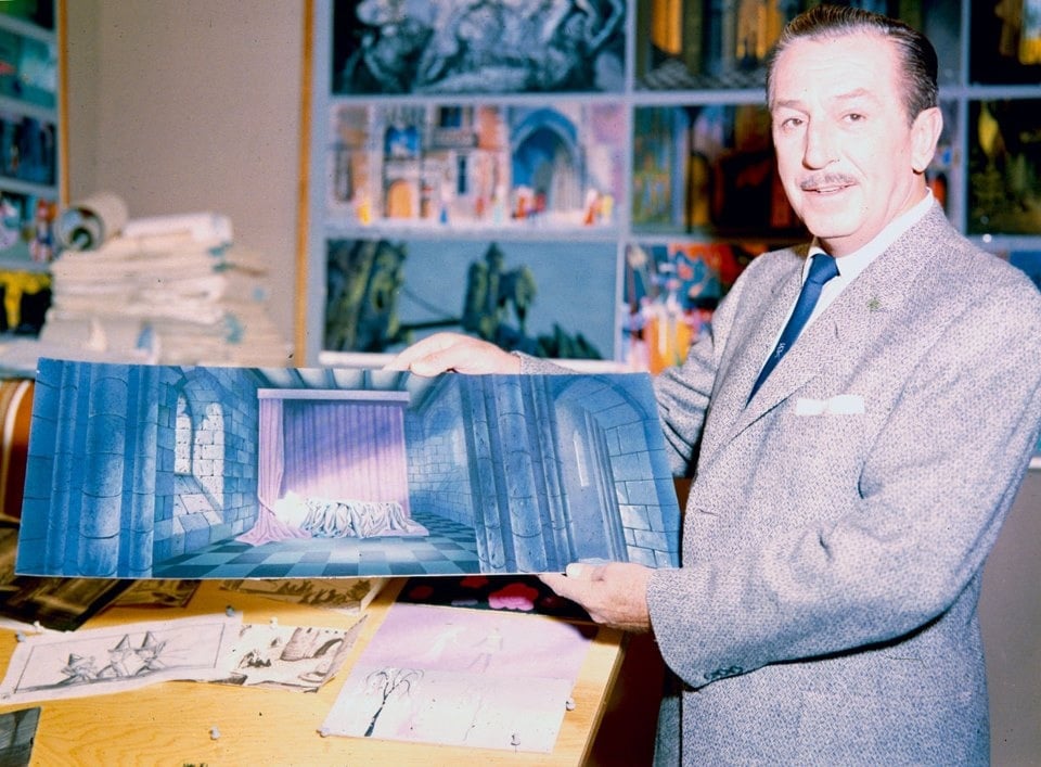 A man in a suit stands in an art studio, holding up a painting of a stage scene with stone walls and a purple curtain. Various drawings and sketches are displayed on the table and wall behind him.