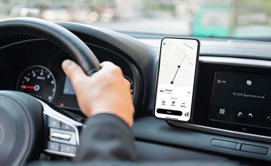 A person drives a car while using a navigation app on a smartphone mounted on the dashboard, showing a map and directions. The driver's left hand is on the steering wheel.