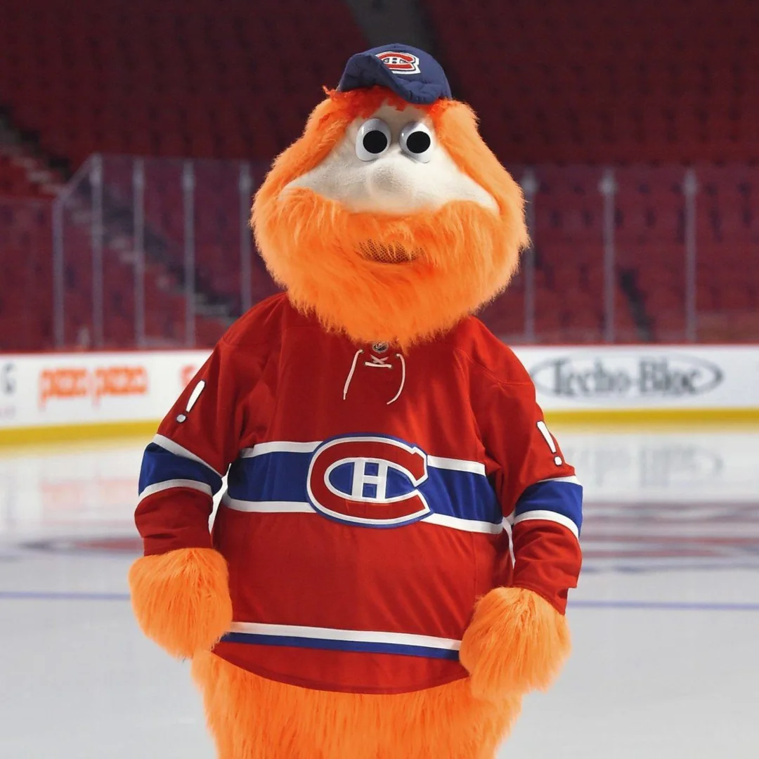 A large orange furry mascot wearing a red Montreal Canadiens hockey jersey and a blue cap stands on an ice rink inside an empty arena.