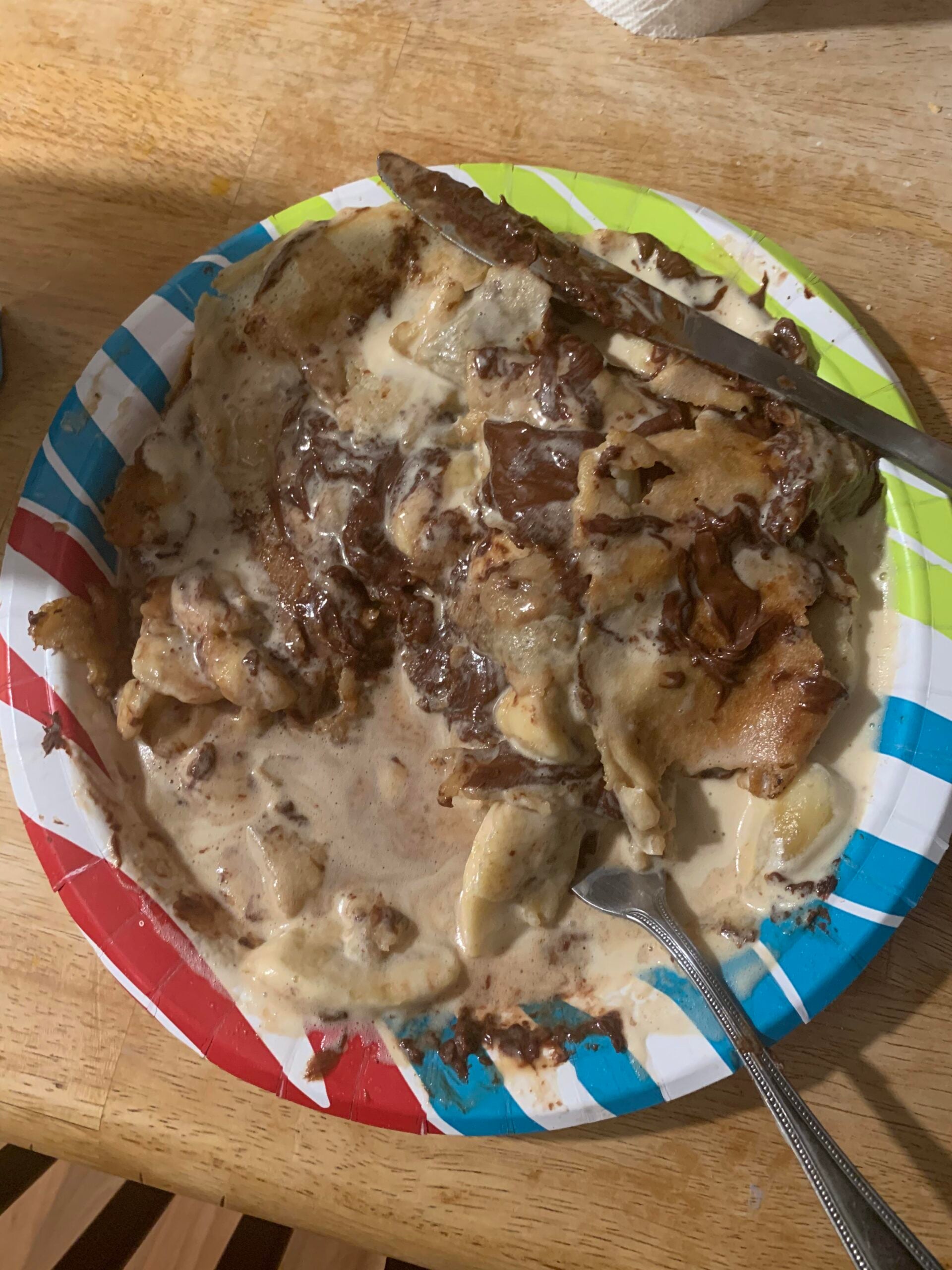 A paper plate with colorful stripes holds a messy serving of bread pudding or French toast covered in creamy sauce, with a fork and knife placed on the plate. The wooden table is visible underneath.
