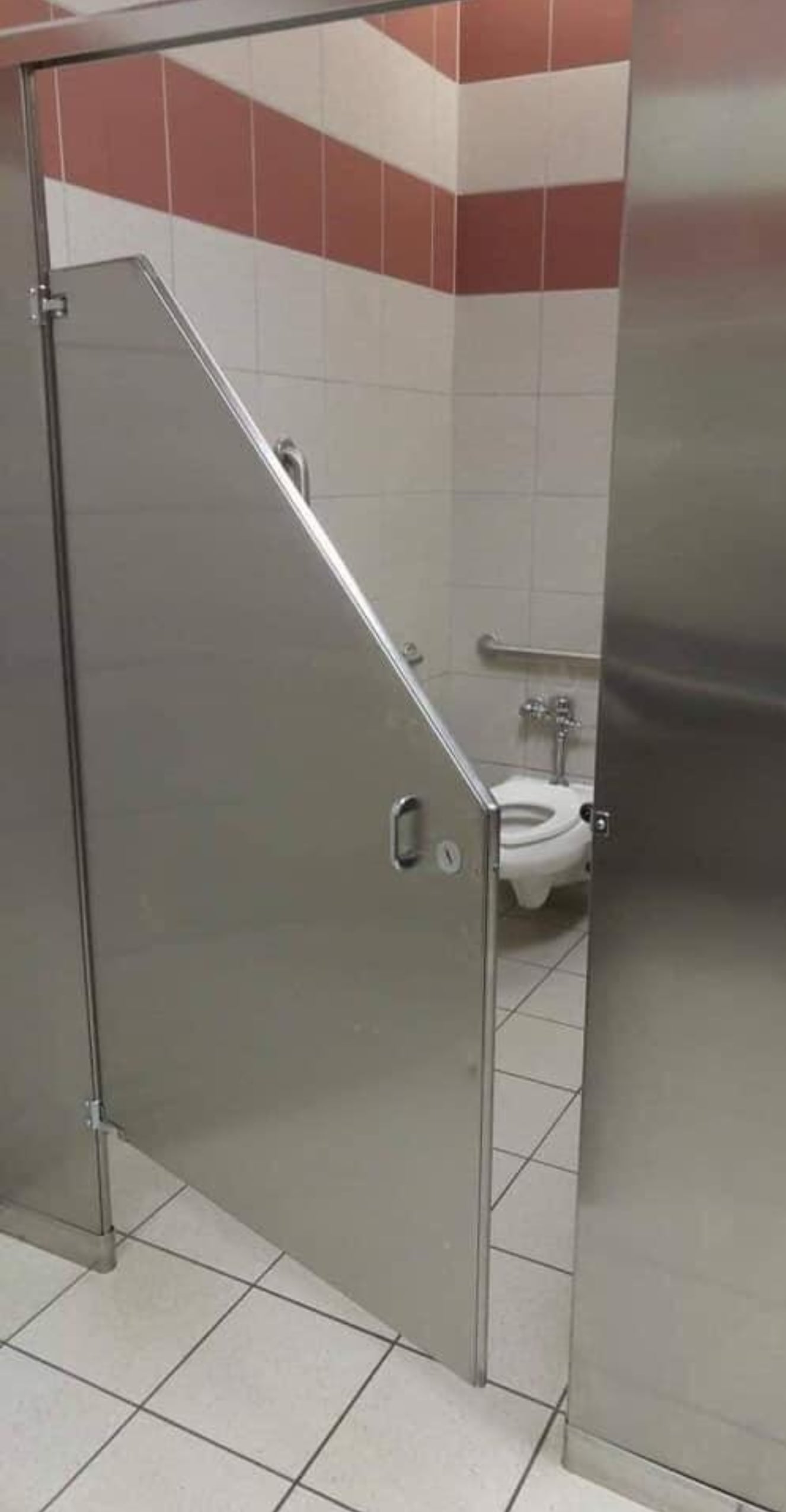 A public restroom stall with a metal door cut at an angle, leaving the toilet inside fully visible from the outside. The walls are tiled white with a red stripe near the ceiling.